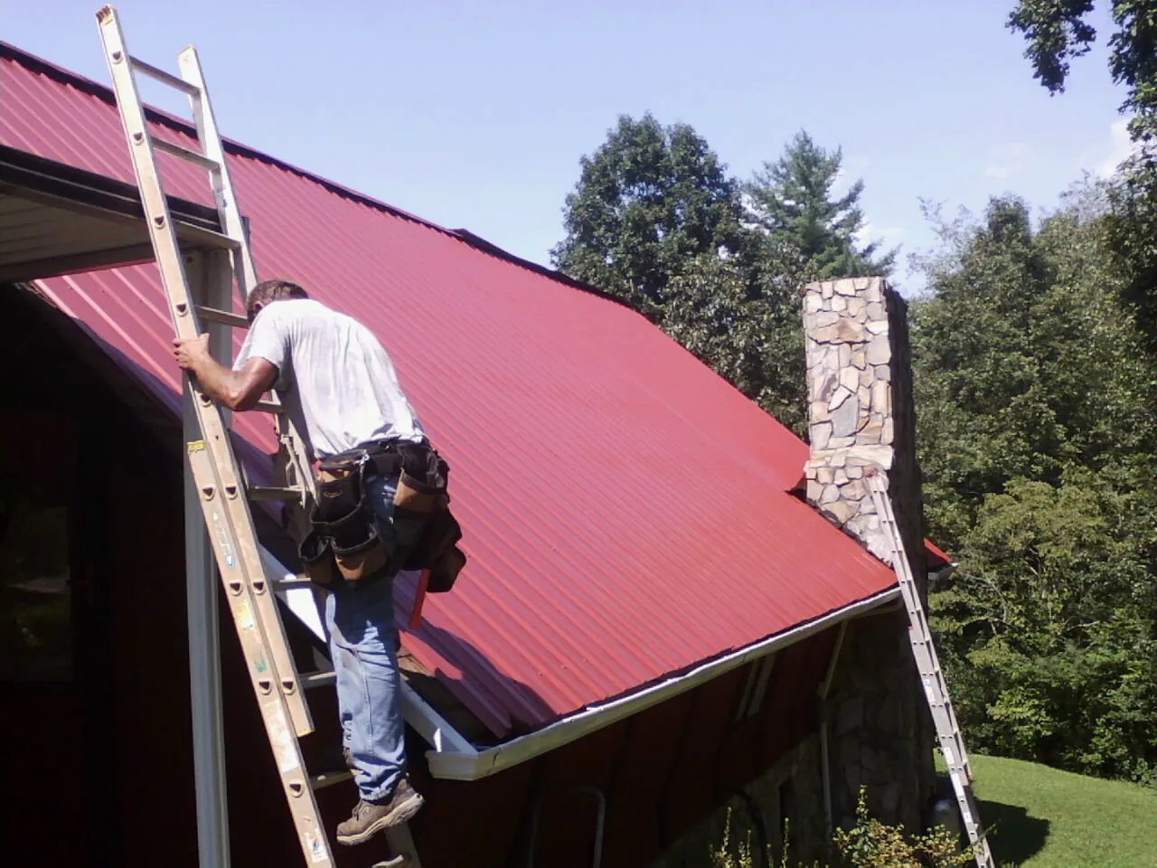 Roofer on a ladder beside a red metal roof and stone chimney — Soffit & Fascia Repair by Roofing Brothers in Westerly